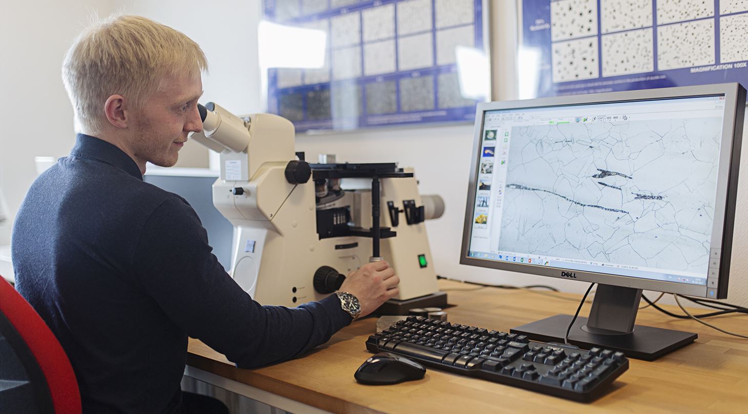 Lyshåret mand studerer emne i mikroskop ved siden af skærm
